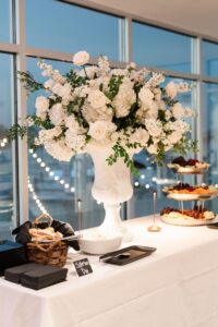 Elegant white floral centerpiece on a table with bread, plates, fruit, cheese; string lights and large windows create a warm wedding vibe.