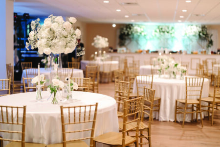 Banquet hall with white-covered round tables, gold chairs, white floral centerpieces, and elegant lighting for Louisiana weddings.