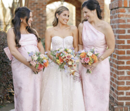 Bride posing with bridesmaids in blush dresses, all holding colorful bouquets in front of brick columns.