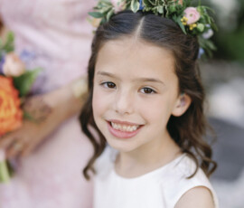 Smiling flower girl wearing a floral crown and white dress, captured in a soft outdoor portrait.