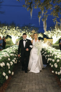 Bride walks down a candlelit garden aisle with her father, surrounded by glowing hedges and white florals at dusk.
