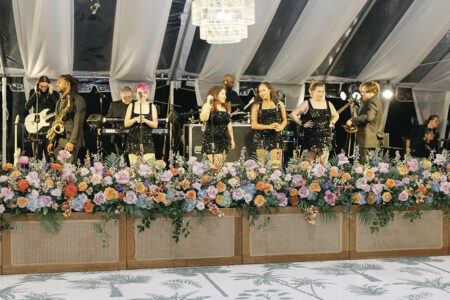 Live wedding band performing on a floral-lined stage beneath a draped tent at the reception.