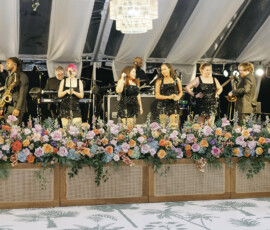 Live wedding band performing on a floral-lined stage beneath a draped tent at the reception.