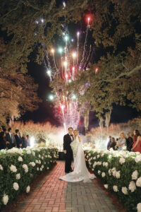 Newlyweds stand together on a candlelit garden aisle as fireworks light up the night sky behind them.
