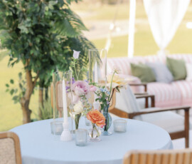 Pastel lounge vignette with a blue cocktail table styled with soft floral arrangements and glass candle holders under a clear-top tent.