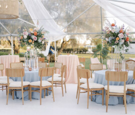 Elegant reception tables featuring pale blue linens, light wood chairs, and lush floral centerpieces beneath a draped clear tent.
