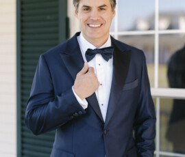 Groom in a classic navy tuxedo adjusting his bow tie, standing outside a white-paneled venue with green shutters.