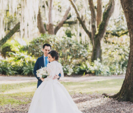 Bride in white gown and groom in blue suit smile together in a sunlit Louisiana garden with trees and hanging moss.