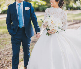 Bride in white gown holding bouquet smiles at groom in blue suit, standing hand-in-hand outdoors amid trees and sunlight.