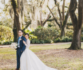 Bride in white gown and groom in blue suit pose in sunlit Louisiana garden with Spanish moss and colorful ornaments.