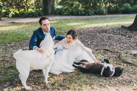 Bride and groom laugh on grass with two dogs during their Louisiana wedding; one dog licks the groom, another gets belly rubs.