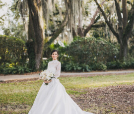 Bride in long white dress smiles with bouquet beneath Spanish moss-draped trees, evoking Louisiana wedding inspiration.