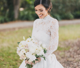 Bride in white lace dress smiles with white bouquet outdoors, green trees and sunlight behind, New Orleans wedding charm.