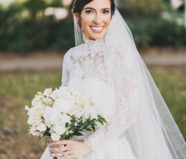 Bride in white lace dress and veil, smiling with bouquet amid lush greenery at a New Orleans wedding.