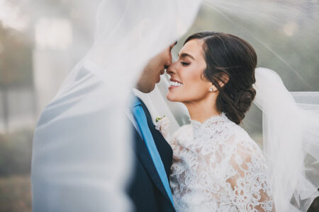 Bride in lace dress and groom in suit smile under veil outdoors at their romantic Gulf South wedding.