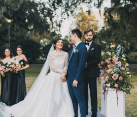 Bride and groom smile at their outdoor Louisiana wedding, joined by bridesmaids in black dresses and an officiant amid flowers.