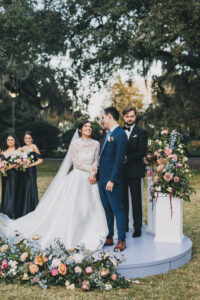 Bride and groom smile at their outdoor Louisiana wedding, joined by bridesmaids in black dresses and an officiant amid flowers.