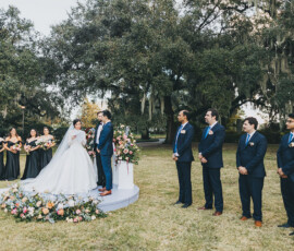 Bride and groom on an outdoor platform with bridesmaids in black, groomsmen in blue, trees and flowers—Gulf South wedding inspiration.