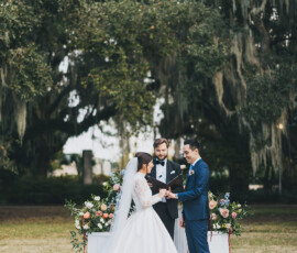 Bride and groom exchange vows before an officiant, surrounded by lush flowers and moss-draped trees at a Gulf South wedding.