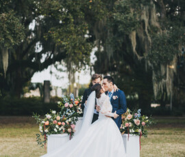 Bride and groom share a kiss at their outdoor Louisiana wedding, with floral arrangements and trees in the background.