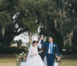 Newlywed couple holds hands and smiles outdoors, surrounded by flowers, greenery, and their officiant under leafy trees.