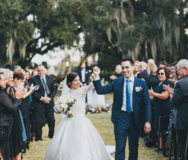 Bride and groom walk hand in hand down an outdoor aisle as guests clap under large trees at a Gulf South wedding.