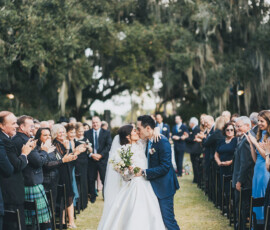 Bride and groom kiss walking down an outdoor aisle, surrounded by cheering wedding guests under large trees.