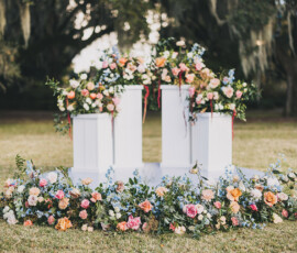 Colorful flower-adorned white pillars on grass, with Spanish moss trees in a lush New Orleans outdoor wedding setting.