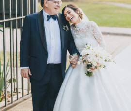 Bride in white gown smiles, holding hands with older man in tuxedo on cobblestone path with flowers, greenery, and buildings.