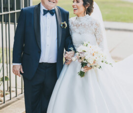 Bride in white gown and veil smiles, holding hands with older man in tuxedo outdoors, greenery background, Gulf South wedding.