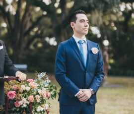 Man in blue suit stands by flower arrangement outdoors, hands clasped, trees behind him at a formal Louisiana event.