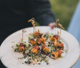 Person in dark clothing serves a white plate with bite-sized appetizers on toothpicks, garnished with microgreens.