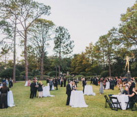 Guests in formalwear mingle by cocktail tables on a lawn with trees, greenery, and a golden statue at a Louisiana wedding.
