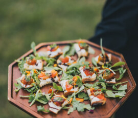 Person in dark clothing holds a wooden tray of bite-sized appetizers with greens and flowers, perfect for wedding inspiration.