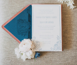 Floral wedding invitation suite with blue envelope, white and blush flowers, and hyacinths on a textured beige surface.