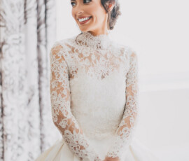 Smiling woman in a white lace wedding dress stands by patterned curtains, hands clasped, inspiring Louisiana wedding style.