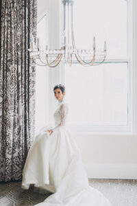 Bride in lace gown stands by a large window under chandelier, holding her dress in an elegant Gulf South wedding setting.