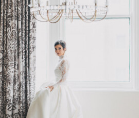 Bride in lace gown stands by a large window under chandelier, holding her dress in an elegant Gulf South wedding setting.