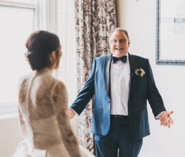 Smiling man in tuxedo gestures to woman in lace wedding dress at joyful New Orleans wedding, bright room in background.