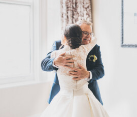 Bride in white lace dress hugs older man in blue suit, both smiling, at a bright Louisiana wedding ceremony.