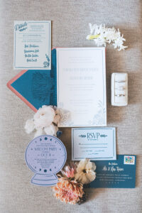 New Orleans wedding stationery flat lay: invites, RSVP, crystal ball card, envelopes, flowers, and ring box on beige background.