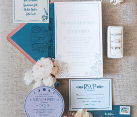 New Orleans wedding stationery flat lay: invites, RSVP, crystal ball card, envelopes, flowers, and ring box on beige background.