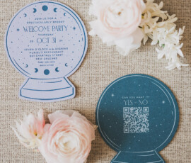 Blue crystal ball-shaped wedding invitations on beige fabric, surrounded by pink and white flowers; one includes a QR code RSVP.