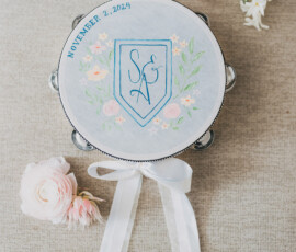 Decorated tambourine with floral art, initials S A, and 11/2/2024, on fabric with pink flowers and white ribbon—Louisiana wedding inspo.