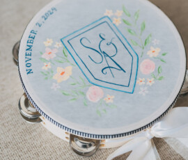 Painted tambourine with floral designs, initials S & N, 11/2/24, on burlap with pink and white flowers—ideal for a New Orleans wedding.
