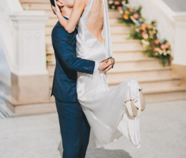 Groom in blue suit lifts and kisses bride in white dress before flower-adorned stairs at New Orleans wedding.