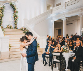 Bride and groom share first dance in an elegant hall with guests, flowers, and a grand staircase—ideal Louisiana wedding inspiration.