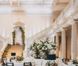Ballroom with round tables, black chairs, white linens, floral accents, and grand staircase—ideal for a Louisiana wedding.
