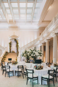 Ballroom with round tables, black chairs, white linens, floral accents, and grand staircase—ideal for a Louisiana wedding.