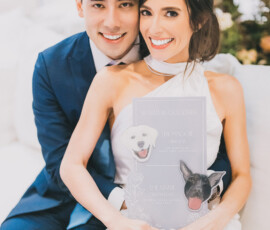 Smiling couple at Gulf South wedding; woman in white dress holds dog-themed cocktail sign, man wears blue suit. Flowers blurred behind.